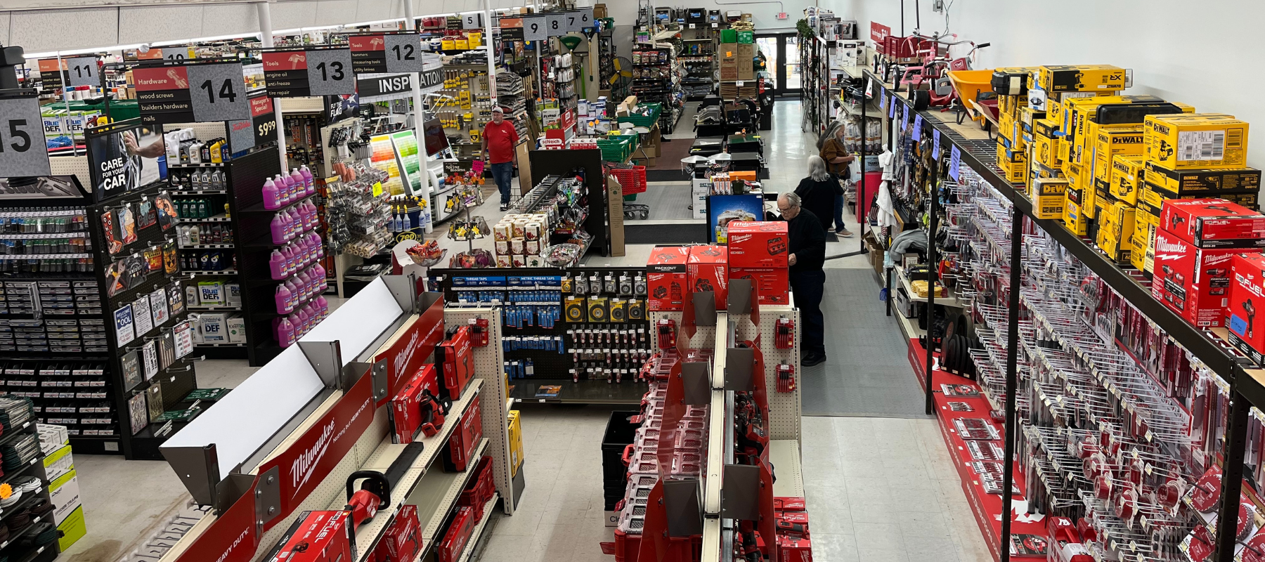 Aerial view of a hardware store aisle, filled with tools and supplies. Brightly colored boxes and shelves line the path, as shoppers browse products.